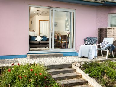 A patio area with a table and chairs outside a pink building with sliding glass doors showing a living room and dining area at Nectan in Mevagissey