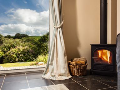 A room with a wood burning stove next to a basket of logs and a window showing a garden at Nectan in Mevagissey