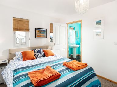 A bedroom with a bed with striped blue blankets and orange towels and pillows and an open door to a turquoise bathroom at Nectan in Mevagissey