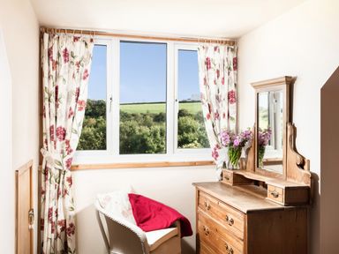 A small room with a wooden dresser with a mirror a chair with cushions and a red blanket and floral curtains by a window at Nectan in Mevagissey