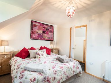 A bedroom with a bed with floral bedspread and red pillows two wooden bedside tables with lamps a textile wall hanging and an open door at Nectan in Mevagissey