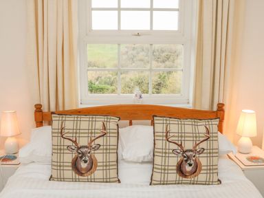 A bed with two deer design pillows and bedside tables with lamps and books by a window with curtains at The Well House Apartment in Watchet