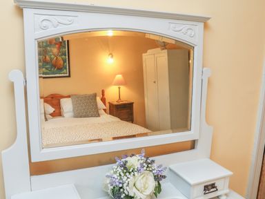 A bedroom reflected in a white mirror showing a bed with pillows a bedside table with a lamp a wardrobe and a framed picture with flowers on a dresser at The Well House Apartment in Watchet