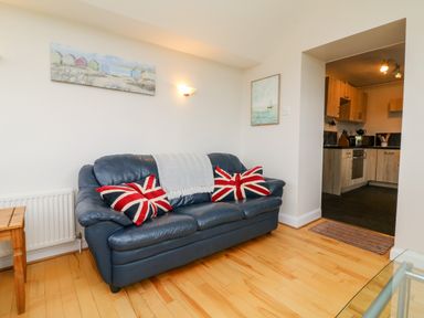 A living room with a blue leather sofa with union jack pillows and a blanket next to a kitchen at The Well House Apartment in Watchet