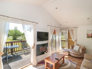 A living room with beige sofas a wooden coffee table a wall-mounted tv and patio doors leading to a deck with chairs and a table at Bramley Lodge in Hewish near Weston-super-Mare