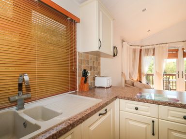 A kitchen with a sink countertop knife block toaster and windows with blinds and curtains at Bramley Lodge in Hewish near Weston-super-Mare