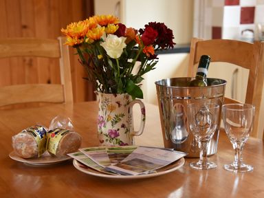 A wooden dining table with a floral jug of flowers a plate with leaflets a plate with wrapped scones a metal ice bucket with a bottle and two glasses at Sycamore Lodge in Nantmel near Rhayader