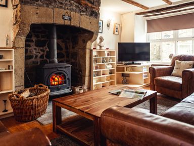 A living room with a stone fireplace and wood burning stove a wooden coffee table leather armchairs a TV and shelves at Bastle House in Haltwhistle near Hadrians Wall