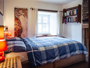 A bedroom with a double bed with blue checkered bedding a window with curtains a wooden bookshelf and a painting on the wall at Bastle House in Haltwhistle near Hadrians Wall