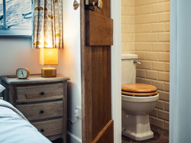 A bedside table with a lamp and clock next to a bed and an open door showing a toilet with a wooden seat and tiled walls at Bastle House Haltwhistle near Hadrians Wall