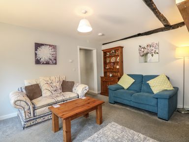 A living room with a patterned sofa, a blue sofa with yellow cushions, a wooden coffee table, a wooden cabinet with decorative plates, and floral paintings at Hadleigh Farm Cottage in Gayton