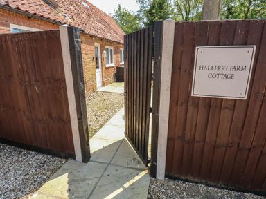 An open wooden gate leading to a pathway beside a brick building at Hadleigh Farm Cottage in Gayton