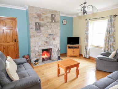 A living room with a stone fireplace a wooden coffee table grey sofas a small wooden TV stand with a television and patterned curtains at Tieve Baun in Ballintrillick near Kinlough County Sligo