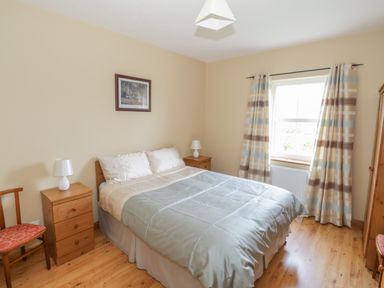 A bedroom with a double bed between two wooden bedside tables with lamps a wooden chair and a window with patterned curtains at Tieve Baun in Ballintrillick near Kinlough County Sligo