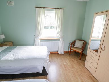 A bedroom with two beds a window with white curtains a wooden chair with a pillow a wardrobe with a mirror and wooden flooring at Tieve Baun in Ballintrillick near Kinlough County Sligo