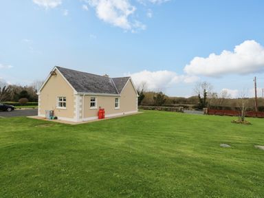 A single-story beige house with a gray roof on a large green lawn with a driveway and trees in the background at Tieve Baun in Ballintrillick near Kinlough County Sligo