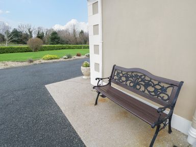 An outdoor bench on a paved area next to a beige wall with a flower pot and greenery in the background at Tieve Baun in Ballintrillick near Kinlough County Sligo