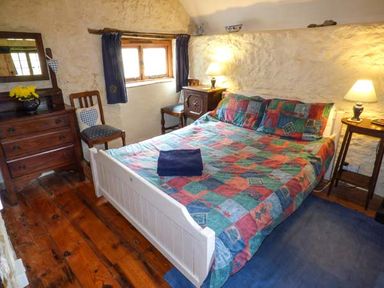 A bedroom with a bed covered with a colorful quilt two bedside tables with lamps a wooden dresser two chairs and a window with blue curtains at The Mill in Bicknoller
