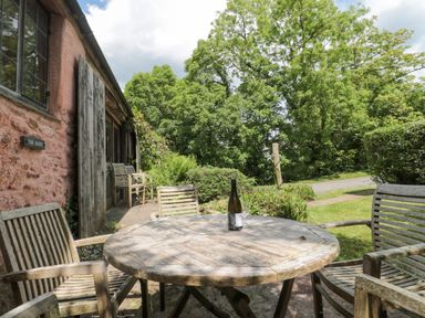 An outdoor patio with a round wooden table and chairs with a bottle on the table next to a rustic building and surrounded by trees at The Barn in Bicknoller