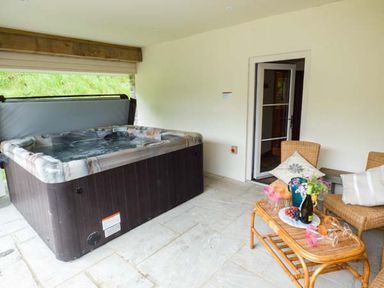 A covered patio with a hot tub and wicker furniture with drinks and flowers at The Old Mill in Dolau near Llandrindod Wells