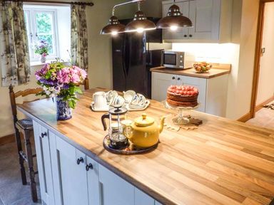A kitchen with a wooden island counter with a teapot and coffee press on a tray a cake on a stand cups and saucers and a vase of flowers at The Old Mill in Dolau near Llandrindod Wells