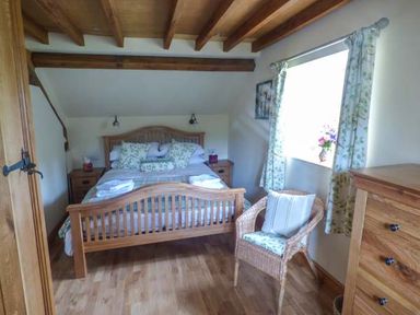 A bedroom with a wooden bed and floral pillows next to a window with patterned curtains and a wicker chair at The Old Mill in Dolau near Llandrindod Wells