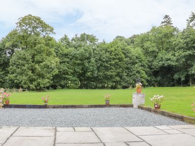 A garden with potted flowers on gravel and grass with trees in the background at The Old Mill in Dolau near Llandrindod Wells