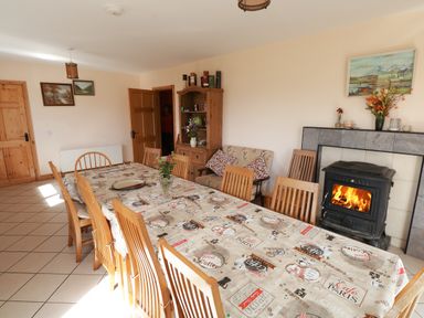 A dining room with a long table covered with a coffee-themed tablecloth surrounded by wooden chairs a wood-burning stove with a fire a wooden cabinet with flowers and framed pictures on the walls at Lough Mask Road Fishing Lodge in Ballinrobe County Mayo