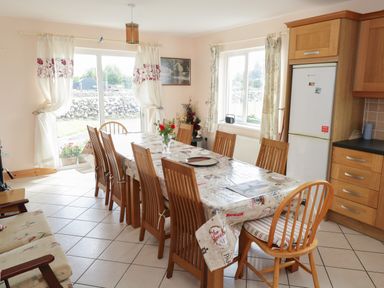 A dining room with a long table surrounded by wooden chairs next to a kitchen with wooden cabinets and a refrigerator at Lough Mask Road Fishing Lodge in Ballinrobe County Mayo