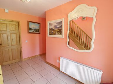 A hallway with orange walls a tiled floor a wooden door two framed paintings a white decorative mirror reflecting a wooden staircase and a white radiator at Lough Mask Road Fishing Lodge in Ballinrobe County Mayo