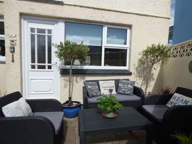 An outdoor seating area with black wicker furniture cushions pillows and potted plants against a beige wall at Spindrift in Porth near Newquay