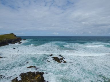 An ocean with waves crashing against rocks and a grassy cliff in the distance at Spindrift in Porth near Newquay