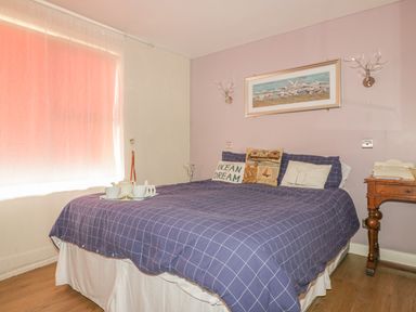 A bedroom with a double bed with a blue checkered bedspread and pillows next to a window with curtains and a wooden bedside table at Spindrift in Porth near Newquay