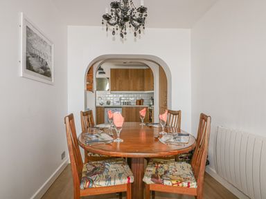 A dining area with a wooden table and six chairs with patterned cushions a chandelier above and an open view into a kitchen at Spindrift in Porth near Newquay