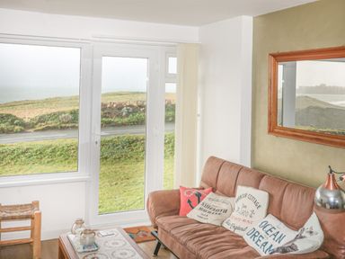 A living room with a brown couch with cushions a wooden coffee table a lamp and large windows with a view of grass and road at Spindrift in Porth near Newquay