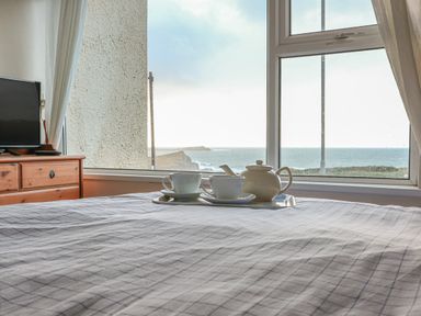 A bedroom with a bed covered in a checkered duvet a tray with a teapot and cups on the bed a wooden dresser with a television and a window showing a sea view at Spindrift in Porth near Newquay
