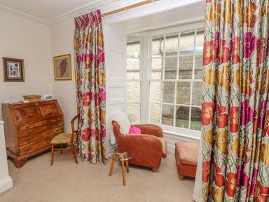 A corner of a room with a wooden desk chair and armchair beside large windows with floral curtains at Berlin House in Tenby
