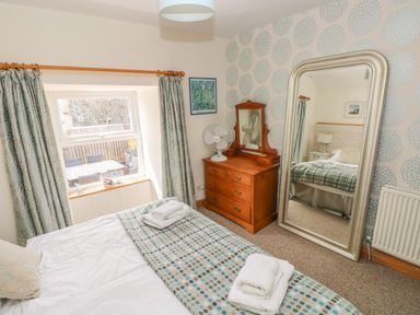 A bedroom with a bed, wooden dresser with mirror, large standing mirror, window with curtains, and patterned wallpaper at Berlin House in Tenby
