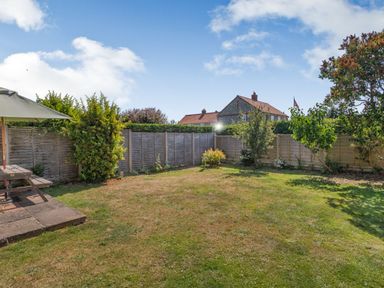 A backyard with grass, wooden fence, bushes, trees, and a picnic table with an umbrella at 38 Dale End in Brancaster Staithe near Burnham Market