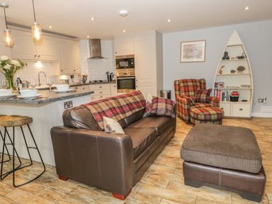 An open-plan kitchen and living room with a brown leather sofa, plaid armchair, ottoman, kitchen island with stools, and a boat-shaped bookshelf at The Old Laundry in Clappersgate near Ambleside