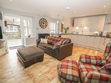 A kitchen and living area with a brown leather sofa, plaid armchair, ottoman, clock on the wall, and white cabinets at The Old Laundry in Clappersgate near Ambleside