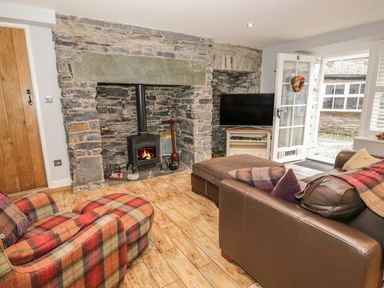 A living room with a stone fireplace a wood stove a plaid armchair a leather sofa a television and an open door at The Old Laundry in Clappersgate near Ambleside