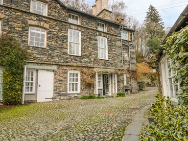 A stone building with white framed windows and doors on a cobblestone path surrounded by greenery at The Old Laundry in Clappersgate near Ambleside