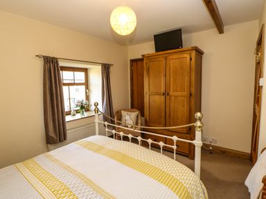 A bedroom with a bed a wooden wardrobe a window with curtains a chair with a cushion and a television on a wardrobe at The Barn Cottage in Steel near Hexham