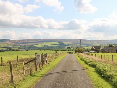 A country road with fences on both sides leading to houses and fields at The Barn Cottage in Steel near Hexham