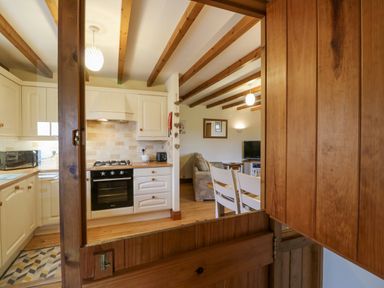 A kitchen with oven and microwave visible through a wooden framed serving hatch into a dining and living area at The Barn Cottage Steel near Hexham