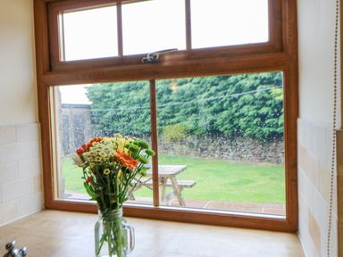 A kitchen window with flowers in a jar on the counter and a garden with a picnic table outside at The Barn Cottage in Steel near Hexham
