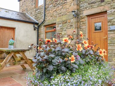 A stone wall house with a wooden door and window a flower bed with orange and purple flowers and a wooden picnic table at The Barn Cottage in Steel near Hexham
