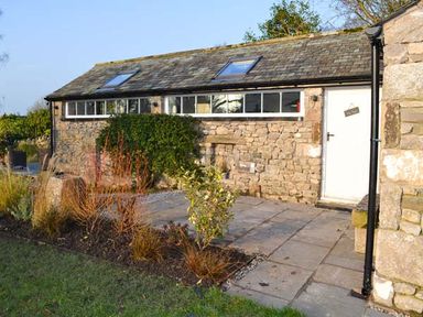 Stone building with a tiled roof and small skylight windows with a white door and garden plants at The Potting Shed in Burton-in-Kendal near Kirkby Lonsdale