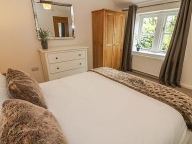 A bedroom with a bed with brown pillows and throw a white dresser with a plant a wooden wardrobe and a window with brown curtains at The Potting Shed in Burton-in-Kendal near Kirkby Lonsdale